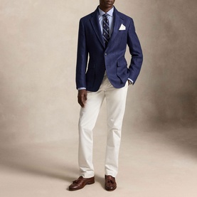Model wearing Ralph Lauren blue reg shirt, styled with navy blazer, striped tie, white trousers and brown loafers.
