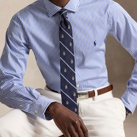 Model wearing Ralph Lauren blue striped regular fit shirt, styled with navy patterned tie and white trousers with brown belt.