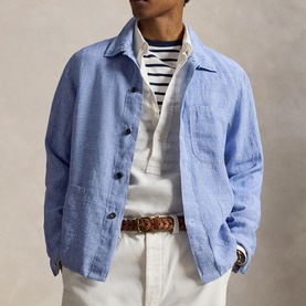 Model wearing light blue linen chore jacket with chest pocket, styled with striped top, white trousers and brown leather belt