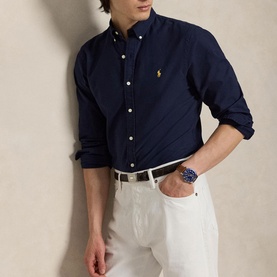 Model wearing Ralph Lauren Slim Shirt in navy with gold pony logo, styled with white trousers and a blue watch, sleeves rolled up.