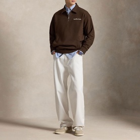Model wearing Ralph Lauren nutmeg brown half-zip sweatshirt with logo, styled with light blue shirt, white trousers and cream trainers.