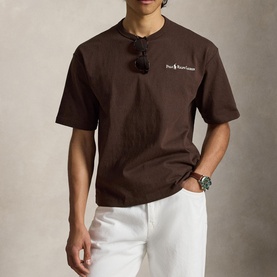 Model wearing Ralph Lauren nutmeg brown crew-neck T-shirt with logo print, styled with white trousers and sunglasses hooked at neckline.