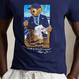 Model wearing Ralph Lauren Newport Navy T-shirt featuring Polo Bear graphic in nautical sailor outfit, seated in wooden chair