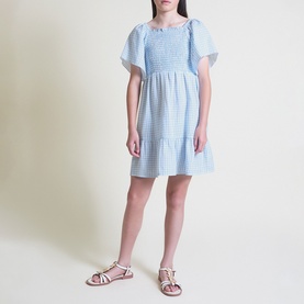 Model wearing Street Monkey blue gingham smocked dress with puff sleeves and tiered skirt, styled with white strappy sandals.