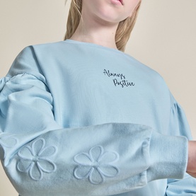 Model wearing Street Monkey Blue Sweat Top with "Always Positive" embroidered text and embossed flower details on sleeve