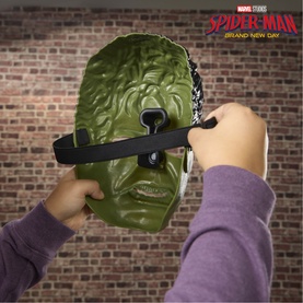 Superheroes Spider-Man Villain Face Mask toy in green, shown being held by child's hands against brick wall background.