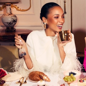 Model wearing white feather-trim jacket, holding gold cup at dining table with pastries and fruit, wearing drop earrings