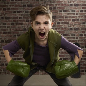 Child wearing Superheroes Hulk Gamma Smash Fists, pair of oversized green foam fists, posed in action stance against brick wall.