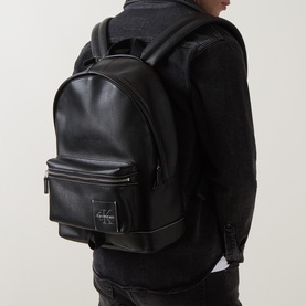 Model carrying Calvin Klein Logo Pebbled Backpack in black faux leather, with logo patch, front zip pocket and adjustable shoulder straps.