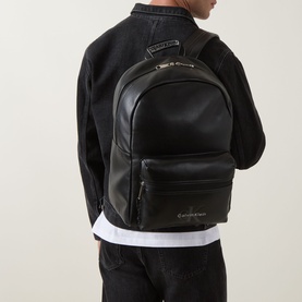 Model carrying Calvin Klein Bold Web Monogram Logo Backpack in black, smooth finish with front zip pocket, gold-tone zips and embroidered logo detail.