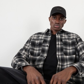 Model with Calvin Klein Logo UV Adjustable Cap in black, curved visor with logo lettering, styled with black T-shirt and plaid overshirt.