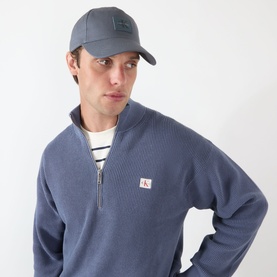 Model with Calvin Klein Rubber Logo Baseball Cap in Turbulence, styled with a blue zip-neck sweatshirt and striped top.