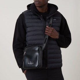 Model carrying Calvin Klein Bold Logo Messenger Bag in black with silver-tone hardware and adjustable webbed strap, styled with black padded gilet.