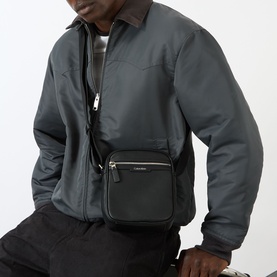 Model carrying Calvin Klein Perforated Reporter Bag in black, crossbody style with silver zip and logo lettering, styled with grey bomber jacket