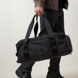 Model carrying Calvin Klein Utility Duffle Bag in black, tonal monogram fabric, logo badge, short handles, styled with black trousers and boots