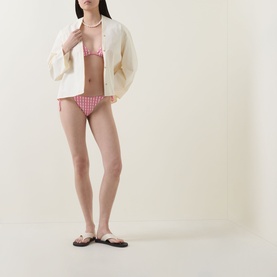 Model wearing Seafolly Bella Gingham Side-Tie Rio Bikini Bottoms in Begonia Pink with matching bikini top, cream oversized shirt and flat sandals.