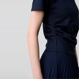Model wearing Claude Pierlot blue short-sleeve top with ruched waist detail, styled with a navy pleated skirt.