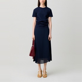 Model wearing Claude Pierlot blue short-sleeve top with ruched waist and navy pleated midi skirt, styled with gold sandals and burgundy bag.