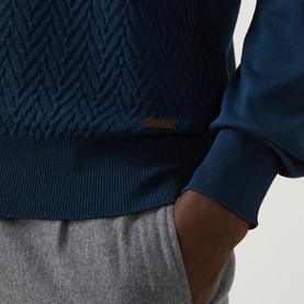 Model wearing Eton Chevron Structure Polo Shirt in mid blue, showing chevron knit texture, ribbed hem, and logo badge detail. Styled with grey