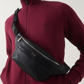 Model carrying Hugo Boss Ray Logo Belt Bag in black pebbled faux leather with silver-toned hardware, worn cross-body over red zip-up top.