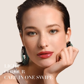 Model with Armani Beauty Prisma Flash Lip Balm, close-up showing glossy rose-tinted lips and dewy skin, wearing dark statement earrings
