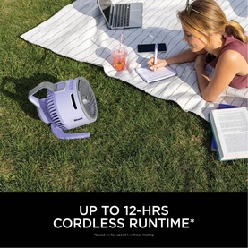 Shark FlexBreeze HydroGo Misting Fan in lavender purple on grass beside woman writing outdoors, text reads up to 12-hrs cordless runtime
