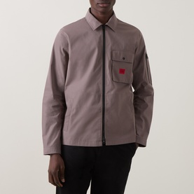 Model wearing taupe zip-through overshirt jacket with chest pocket and small red logo patch, styled with black trousers