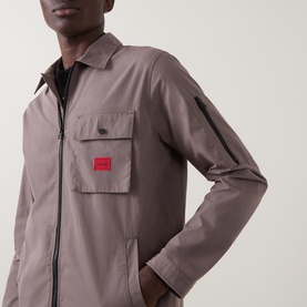 Model wearing a taupe zip-through overshirt jacket with chest pocket and red logo patch, layered over a dark top.