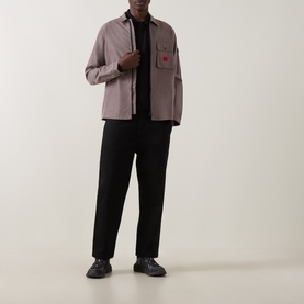 Model wearing a dusty pink overshirt with chest pockets and red logo detail, styled with black trousers and black trainers