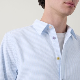 Model wearing Paul Smith blue cotton casual shirt with white stripe and multicoloured buttons, styled with a white crew-neck tee underneath.