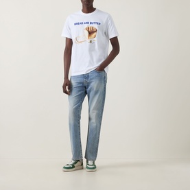 Model wearing Paul Smith Bread & Butter Logo T-Shirt in white with bread graphic print, styled with light wash straight-leg jeans and green trainers.