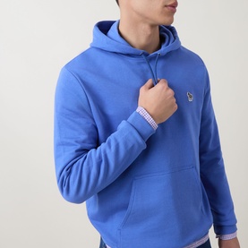 Model wearing Paul Smith blue cotton hoodie with kangaroo pocket and small logo badge on chest, styled with a striped shirt underneath.