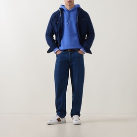 Model wearing Paul Smith blue cotton hoodie, styled with navy overshirt, dark blue jeans and white trainers with stripe detail.
