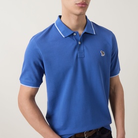Model wearing Paul Smith blue cotton polo shirt with white tipped collar and cuffs, featuring embroidered zebra logo on chest.