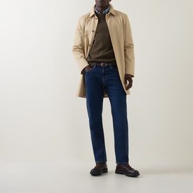 Model wearing Paul Smith blue straight-leg jeans, styled with a beige mac coat, brown knit, and dark leather loafers.