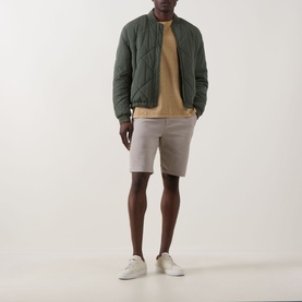 Model wearing dark green quilted bomber jacket, styled with tan sweatshirt, grey shorts, and white trainers