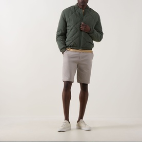 Model wearing green quilted bomber jacket styled with light grey shorts, tan belt, and white trainers