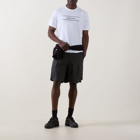 Model wearing Calvin Klein 20S SPW Graphic Logo T-Shirt in Bright White with black cargo shorts, belt bag, and chunky trainers.