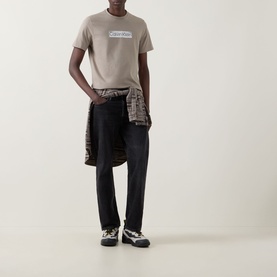 Model wearing Calvin Klein 30s Logo T-Shirt in Roast Cashew, styled with black jeans, plaid shirt tied at waist and trainers.