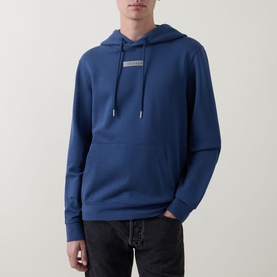 Model wearing Calvin Klein dark denim blue pullover hoodie with small logo patch, kangaroo pocket, and drawstring hood, styled with black jeans