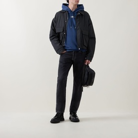 Model wearing Calvin Klein Dark Denim hoodie, styled with black zip jacket, dark jeans, and carrying a black bag.