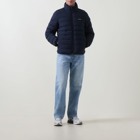 Model wearing Calvin Klein Logo Padded Jacket in Dark Sapphire, quilted funnel neck design, styled with light blue jeans and white trainers.