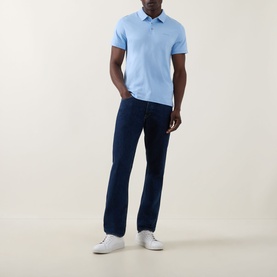 Model wearing Calvin Klein Badge Logo Polo Shirt in Placid Blue, short sleeves, three-button placket, styled with dark navy jeans and white trainers.