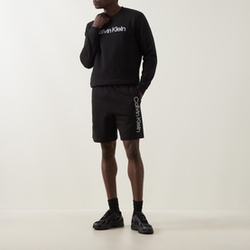 Model wearing Calvin Klein black cotton shorts with logo side print, styled with matching black sweatshirt and black trainers.