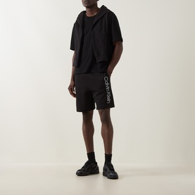 Model wearing Calvin Klein black cotton shorts with logo print, styled with a black short-sleeve hoodie and black trainers.