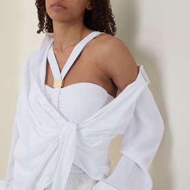 Model wearing white off-shoulder twisted front top with gold-tone necklace detail, styled with a white shirt draped over shoulders