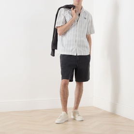 Model wearing Fred Perry Seersucker Check Shirt in Ecru with dark shorts and white trainers, jacket draped over shoulder.