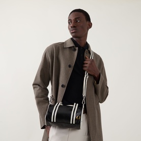 Model carrying Fred Perry Barrel Mini Duffle Bag in Black/Ecru, worn crossbody with white stripe strap, styled with houndstooth coat