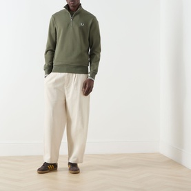 Model wearing Fred Perry Half Zip sweat in Laurel Wreath Green, styled with cream wide-leg trousers and blue striped trainers.