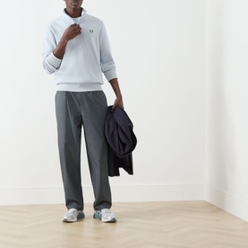 Model wearing Fred Perry Laurel Wreath Half-Zip Sweatshirt in Dusky Blue, styled with grey trousers and white trainers, carrying navy jacket.